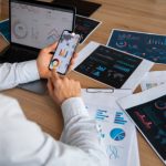 Uomo d'affari analizza dati finanziari su strumenti digitali gratuiti utilizzando smartphone e laptop, circondato da grafici e report stampati su un tavolo di lavoro.