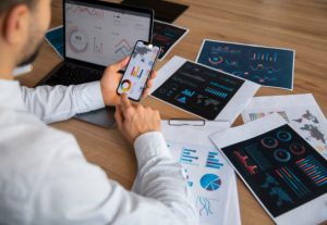 Uomo d'affari analizza dati finanziari su strumenti digitali gratuiti utilizzando smartphone e laptop, circondato da grafici e report stampati su un tavolo di lavoro.
