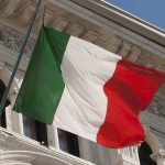 Bandiera dell’Italia che sventola davanti a un edificio storico con architettura classica; l’immagine simboleggia l’identità nazionale ed è spesso associata a istituzioni ufficiali e titoli del governo italiano.