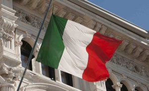 Bandiera dell’Italia che sventola davanti a un edificio storico con architettura classica; l’immagine simboleggia l’identità nazionale ed è spesso associata a istituzioni ufficiali e titoli del governo italiano.